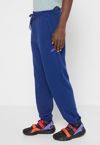 Granatowe dresy z miękkiego materiału z elastycznym pasem i ściągaczami na kostkach; na udzie znajduje się subtelne logo. W zestawie z czarnymi sandałami.