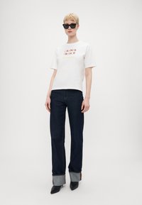 Hvit t-skjorte med rød og gul tekst som sier "CANNES FRANCE Biensur," kombinert med mørkeblå denimjeans med oppvikede bukseben og svarte hæler.