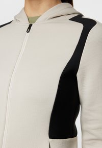 Ljusgrå zip-up hoodie med texturerad tyg, svarta sidodetaljer och en åtsittande passform. Inkluderar en huva och framfickage med dragkedja.