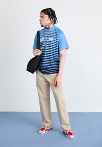 Polo à manches courtes bleu avec des rayures horizontales, pantalon kaki et baskets rouges/grises ; portant un petit sac noir.