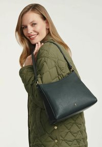 Czarna skórzana torebka o gładkiej fakturze, wyraźnym kształcie i pojedynczym regulowanym pasku, zestawiona z zieloną pikowaną kurtką.