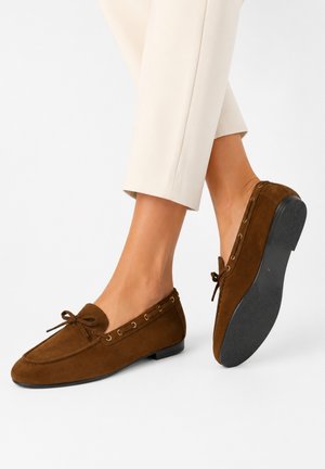 Brune ruskind-loafers med sløjfedetalje, båret med beige afkortede bukser, der viser side- og såludsigter på lys baggrund.
