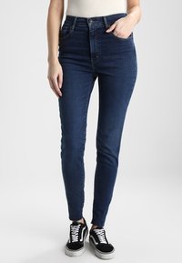 Engpassjeans aus dunkelblauem Denim; hohe Taille mit zwei Vorderaschen; ausgefranste Akzente in der Nähe des Knöchels; kombiniert mit schwarzen Sneakers.