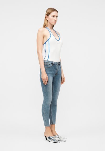 Γυναίκα που στέκεται, φορώντας λευκό τοπ με λαιμόκοψη halter και μπλε περιθώριο, στενά ανοιχτόγάλαζα τζιν και ασημί μυτερά mule με τακούνι σε λευκό φόντο.