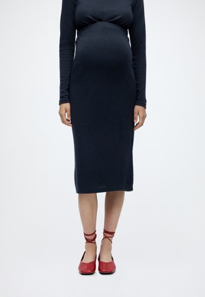 Femme enceinte portant une robe midi noire moulante à manches longues et des chaussures plates rouges à lacets, debout sur un fond blanc.