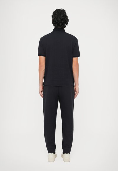 Fekete galléros póló texturált anyagból, rövid ujjú; hozzáillő fekete nadrággal párosítva. A modell háttal áll, bézs tornacipőt visel.