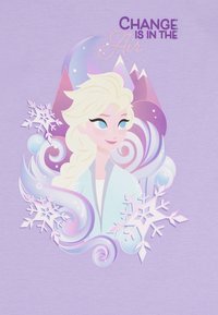 Camisa de color púrpura claro con un personaje estilizado que tiene el pelo rubio, copos de nieve, diseños en espiral y el texto "El cambio está en el aire."