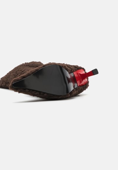Zapatilla de casa de pelaje marrón con una suela negra brillante y un acento rojo en el talón. Presenta una forma puntiaguda y un exterior texturizado.