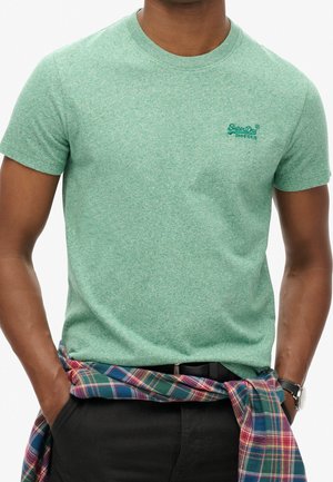 Man draagt een groen Superdry T-shirt met een geruit overhemd om zijn middel gestrikt en een polshorloge om zijn linkerpols.