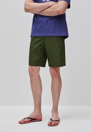 Mężczyzna stoi ze skrzyżowanymi ramionami, ubrany w oliwkowe szorty, granatowy t-shirt oraz czarne i pomarańczowe klapki na jednolitym jasnym tle.