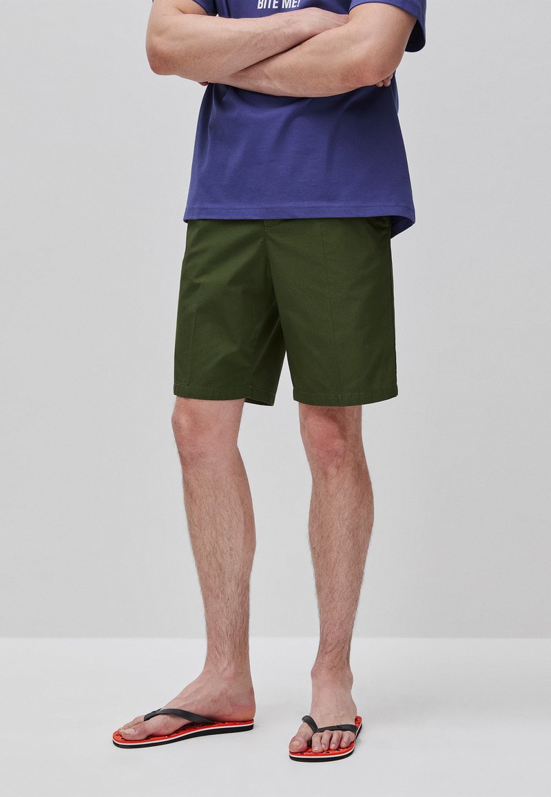 Mann steht mit verschränkten Armen, trägt olivgrüne Shorts, ein dunkelblaues T-Shirt und schwarze-orange Flip-Flops auf einem einfarbigen hellen Hintergrund.