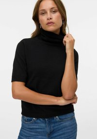 Černý krátký rukáv turtleneck svetr s přiléhavým střihem. Měkká textura, žebrovaný límec a střih do boků v kombinaci s modrými džíny.