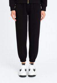 Pantaloni della tuta neri con caviglie affusolate, caratterizzati da una texture liscia e pieghe frontali, abbinati a sneakers bianche con accenti neri.