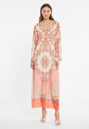 Woman wearing a long-sleeve, floor-length dress with intricate pink, orange, and beige paisley patterns, paired with white strappy heels.