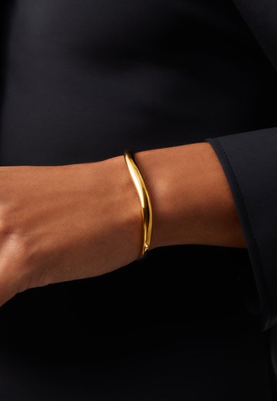 Καρπό με λείο, λαμπερό χρυσό βραχιόλι που φοριέται πάνω από μαύρο ρούχο με μακριά μανίκια σε σκουρόχρωμο φόντο.