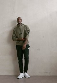 Felpa verde oliva con colletto alto e dettagli neri abbinata a pantaloni jogger verde scuro. Presenta loghi Adidas bianchi e sneaker leggere.