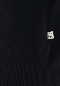 Schwarzer gestrickter Stoff mit geripptem Saum und einem kleinen weißen Etikett, das ein kreisförmiges Symbol am rechten Rand zeigt.