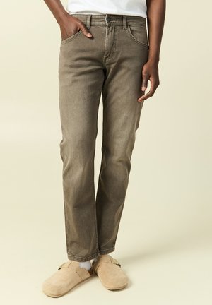 Persona con jeans marrones de pierna recta, zapatos beige sin cordones con hebillas y camisa blanca, de pie con una mano en el bolsillo.