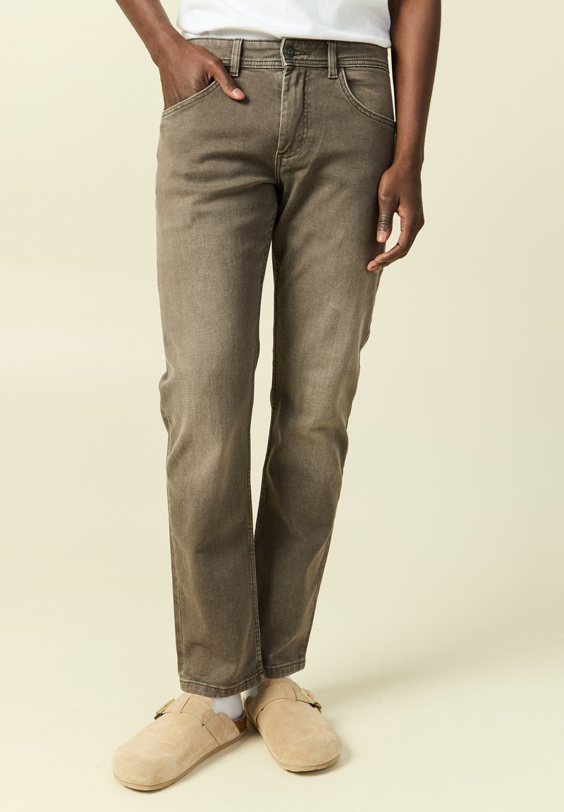 Osoba ubrana w brązowe proste dżinsy, beżowe wsuwane buty z klamrami oraz białą koszulę, stojąca z jedną ręką w kieszeni.