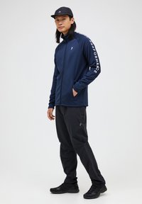 Marinegrøn zip-up jakke med hvid "PERFORMANCE" tekst på ærmet, parret med sorte bukser og sneakers, båret af en person, der står i en neutral stilling.
