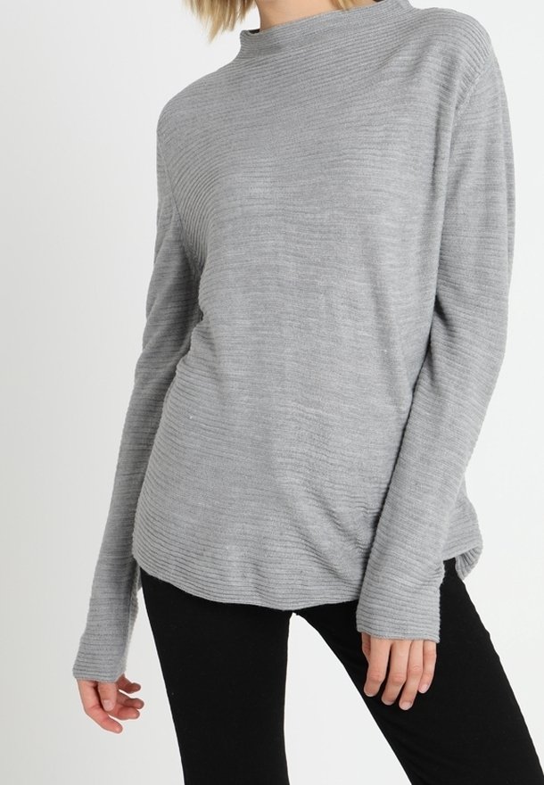 Femme portant un pull à côtes gris clair à manches longues associé à un pantalon noir, debout devant un fond blanc uni.