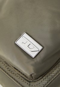Šviesiai žalia nailono rankinė su lygiu paviršiumi, ant kurios priekio yra sidabrinė stačiakampė detalių logotipo akcentas.