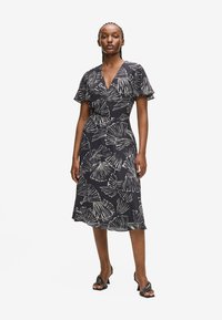 Robe noire avec motif de feuilles blanches, manches courtes volantées, décolleté en V et jupe fluide jusqu'aux genoux. Associée à des sandales à talons noires.