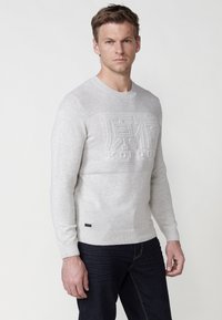 Lichtgrijze gebreide trui met een textuurpatroon op de borst, met een verhoogd logo en geribde manchetten en zoom. Gestyled met donkere jeans.