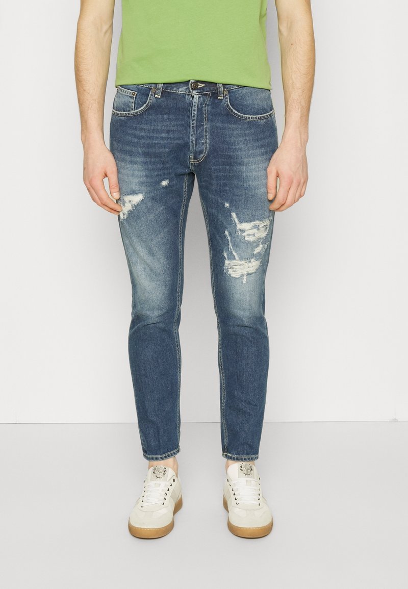 Homme portant un jean bleu déchiré, des baskets blanches et une chemise verte, debout devant un fond blanc uni.