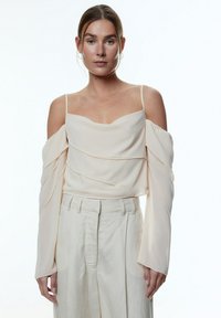 Helle beige schulterfreie Bluse mit geschichteter Textur und langen, fließenden Ärmeln, kombiniert mit cremefarbenen, weit geschnittenen Hosen.