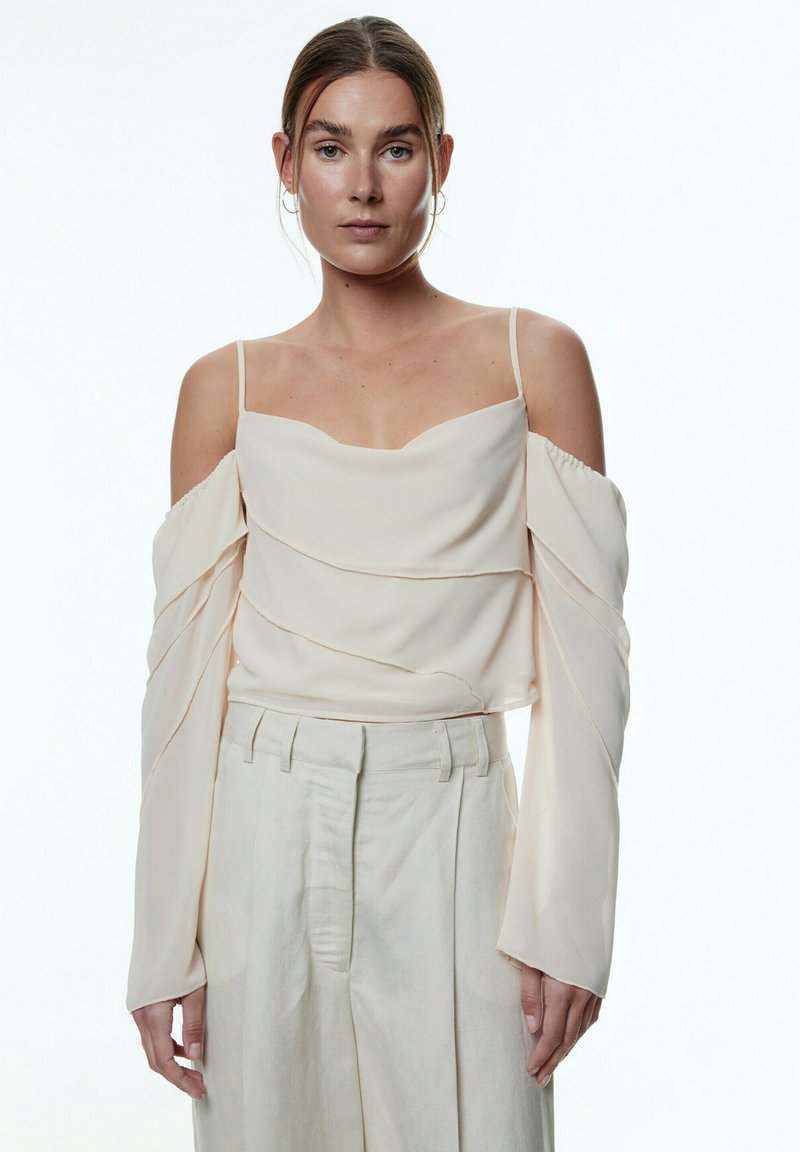 Helle beige schulterfreie Bluse mit geschichteter Textur und langen, fließenden Ärmeln, kombiniert mit cremefarbenen, weit geschnittenen Hosen.