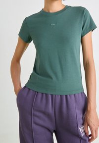 Camiseta verde ajustada de manga corta y cuello redondo, con un pequeño logotipo de Nike en un tono más oscuro, combinada con pantalones de chándal morados.