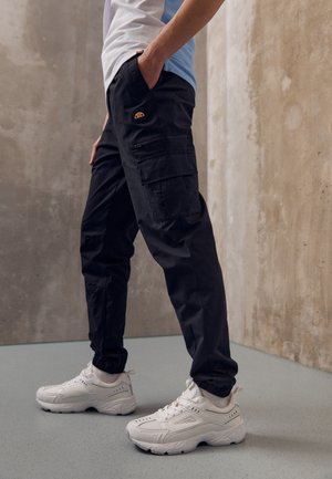 Personne portant un pantalon cargo noir avec un logo sur la poche et des chaussures de sport blanches, debout contre un mur beige texturé.