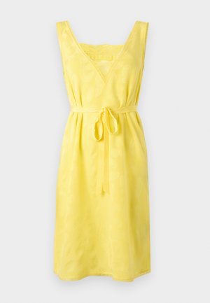 Žena stojaca s rukami skríženými za chrbtom, oblečená v žltých šatách bez rukávov so zaväzovaním v páse a v čiernych čižmách po lýtka na bielom pozadí.