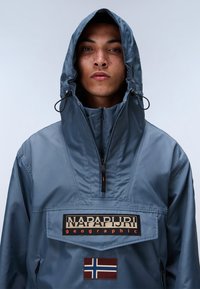 Blue waterproof parka with a high neck, adjustable hood, zippered closure, and a front patch featuring logo and flag details. Smooth texture.
