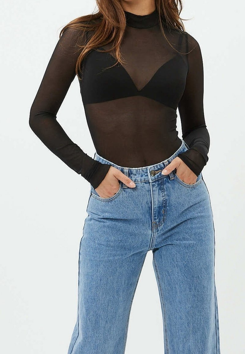 Femme portant un haut noir transparent à manches longues avec un soutien-gorge noir en dessous et un jean taille haute bleu clair, mains dans les poches.