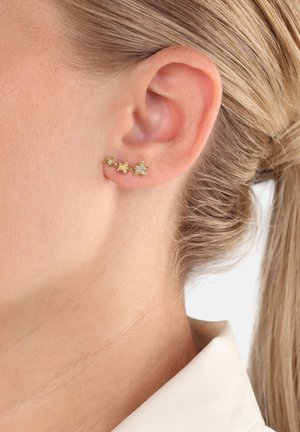 Ohrringe in Goldfarbe mit drei Sternformen, zwei in Gold und eine mit einem Kristallakzent, die am Ohrläppchen hängen.