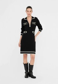 Črna pletena obleka z zadrgo na izrezu, belim robom in dekorativnimi zlatimi poudarki. Ima dolge rokave in oprijet silhueto.