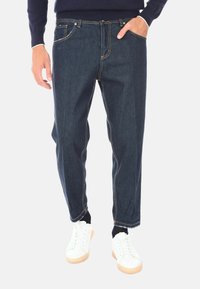 Jeans in denim blu scuro con vestibilità rilassata, caratterizzati da cuciture gialle, cinque tasche e un design a gamba dritta, abbinati a sneaker bianche.