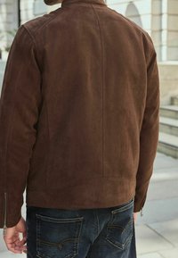 Giacca in suede marrone con colletto rialzato, polsini con zip e texture morbida. Indossata sopra jeans in denim scuro, che mettono in risalto un design minimalista.