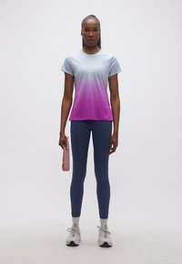 Γυναίκα που στέκεται φορώντας ένα αθλητικό πουκάμισο με βαθμιδωτό χρώμα, σκούρο μπλε κολάν, λευκά αθλητικά παπούτσια, κρατώντας μια ροζ ζώνη αντίστασης στο πλάι της.