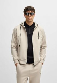 Hoodie zippé beige avec des accents noirs, doté de cordons de serrage et d'un logo sur la poitrine gauche, associé à un polo noir et un pantalon assorti.