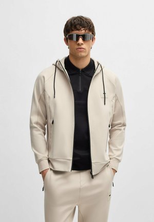 Felpa beige con zip e dettagli neri, con cordini e logo sul lato sinistro del petto, abbinata a un polo nera e pantaloni coordinati.