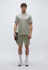 Αθλητικό πουκάμισο και σορτς σε ελιά πράσινο, κοντά μανίκια, μαλακό ύφασμα, μονόχρωμο, σε συνδυασμό με λευκές κάλτσες και αθλητικά παπούτσια, κρατώντας ένα μαύρο μπουκάλι νερού.