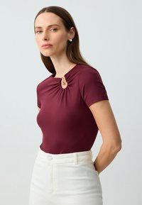 Burgundowa bluzka z krótkim rękawem, z zebranym dekoltem i złotym akcentem. Noszona z białymi spodniami o wysokim stanie, charakteryzującymi się gładką teksturą.