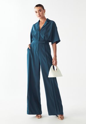 Tiefer, weiten Bein Overall in Teal mit kragenförmigem Ausschnitt und kurzen Ärmeln, ausgestattet mit einem taillierten Gürtel. Begleitet von einer weißen Handtasche mit einem runden Griff.