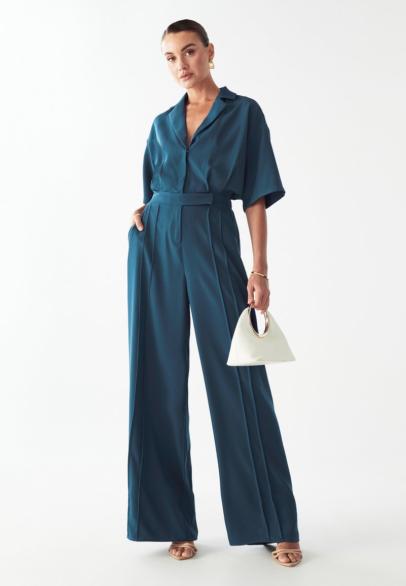 Mono de pierna ancha en tono verde azulado, con cuello tipo camisa y mangas cortas, que presenta una cintura ceñida con cinturón. Acompañado de un bolso blanco con un asa circular.