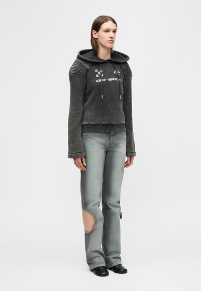 Νεαρή γυναίκα που φοράει σκούρο γκρι φούτερ με κουκούλα και λευκό κείμενο, καθώς και ξεβαμμένο γκρι τζιν με μεγάλο σκίσιμο σε ένα γόνατο, στέκεται μπροστά σε απλό φόντο.