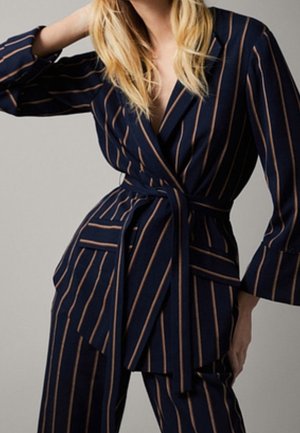 Femme portant un costume bleu marine avec de fines rayures verticales marron et une taille ceinturée, posant avec un bras levé et l’autre sur la hanche.