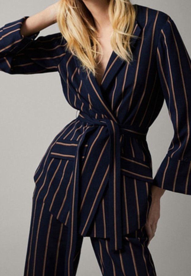 Femme portant un costume bleu marine avec de fines rayures verticales marron et une taille ceinturée, posant avec un bras levé et l’autre sur la hanche.